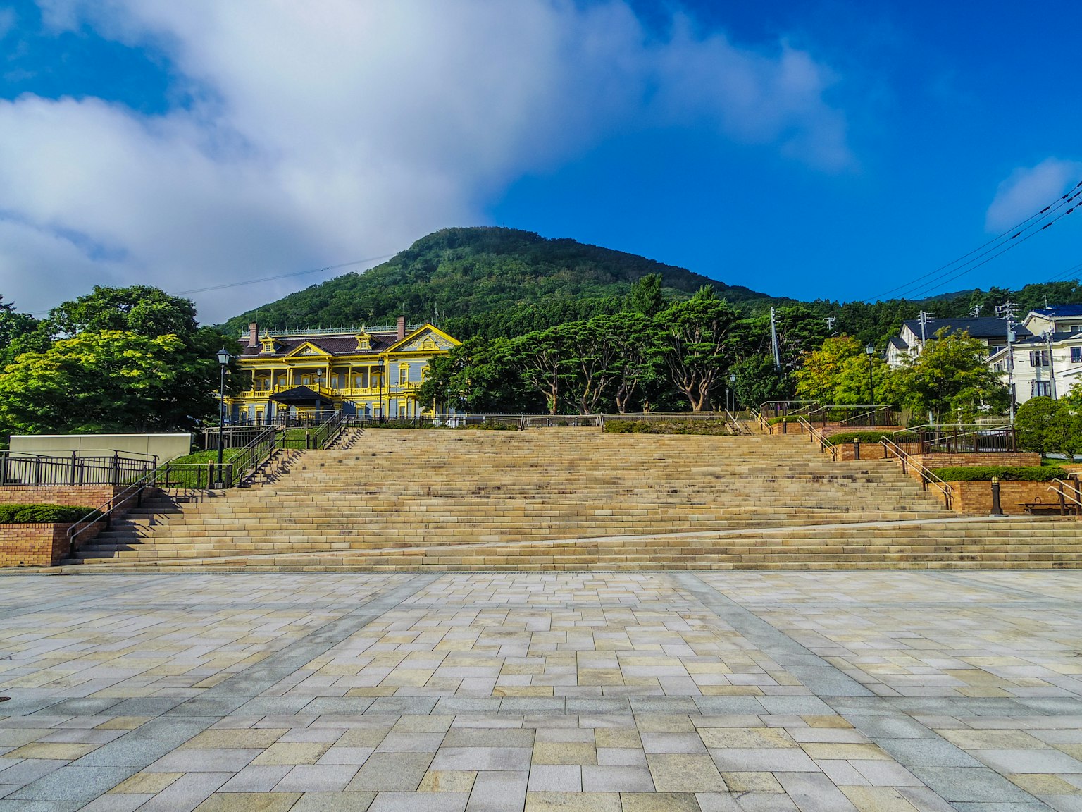 Scenic view featuring a stone-paved plaza and a yellow building surrounded by greenery under a blue sky