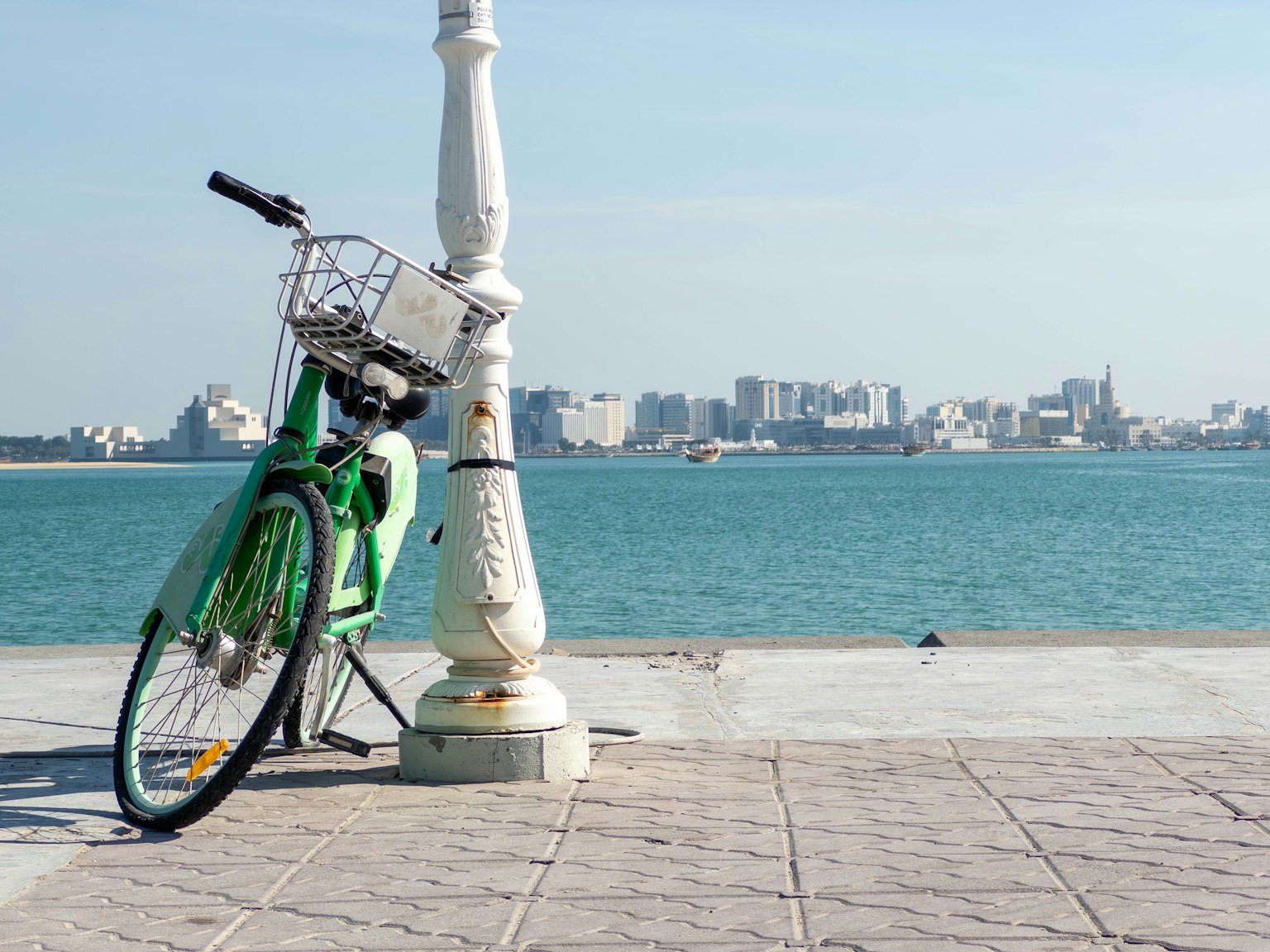 Grünes Fahrrad lehnt an einem weißen Pfosten mit blauem Meer und Stadtansicht im Hintergrund