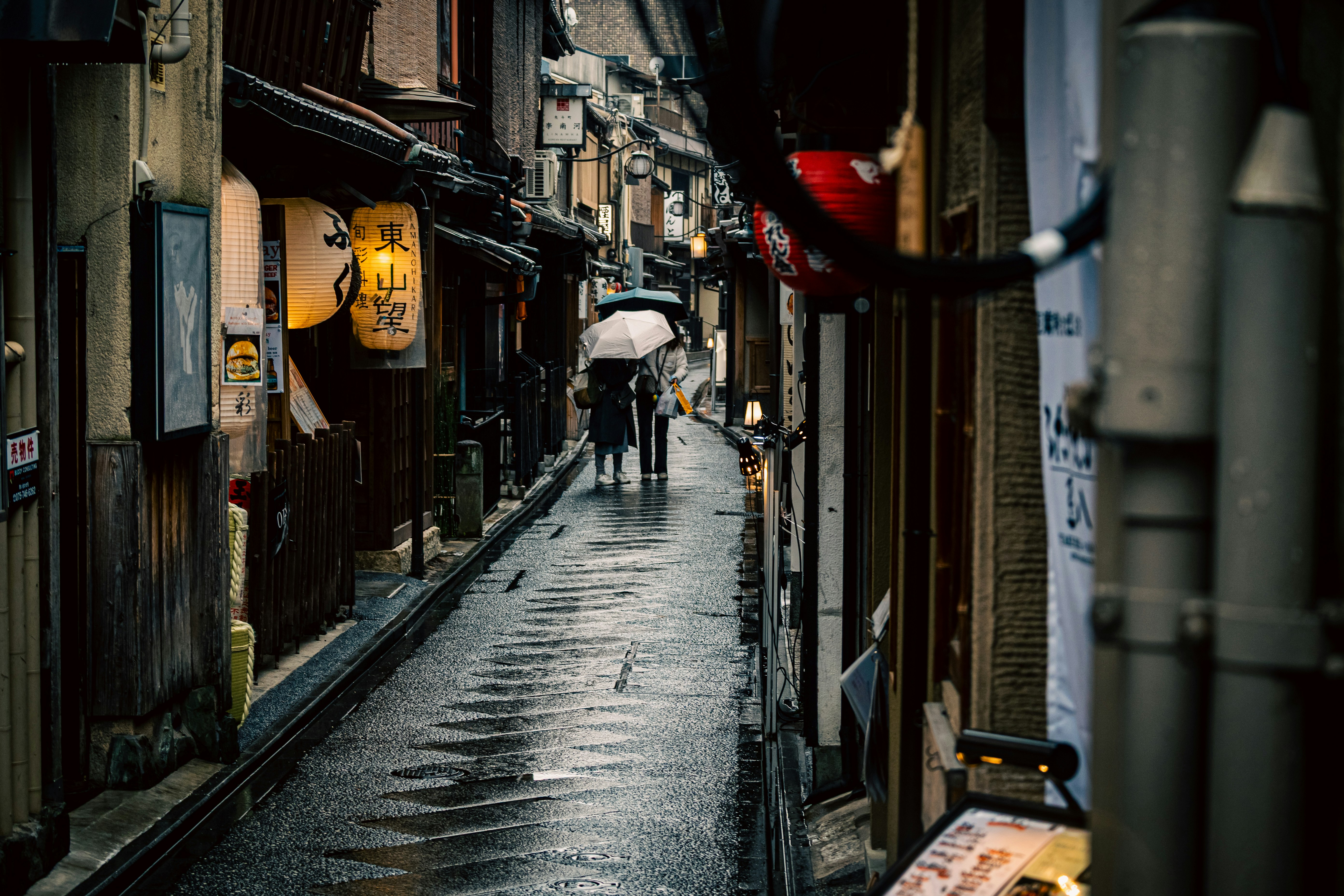 雨の路地に並ぶ店先と傘の人影