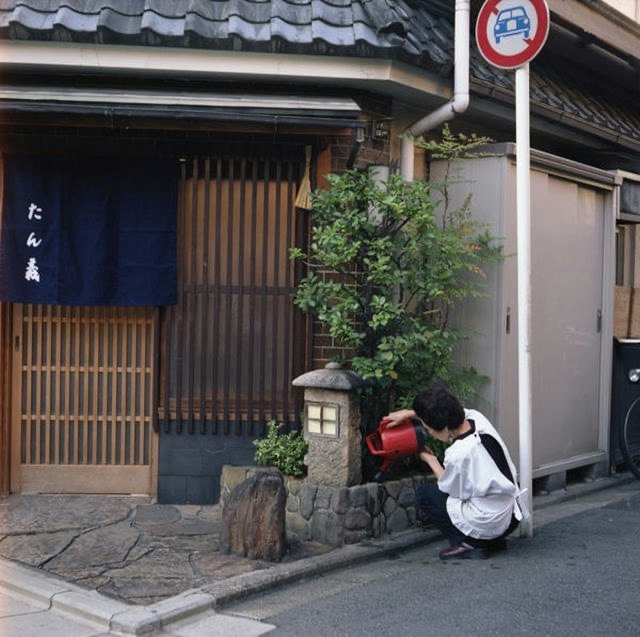 京都の路地にある町家の軒先と石畳、赤いポストの前でしゃがむ人物