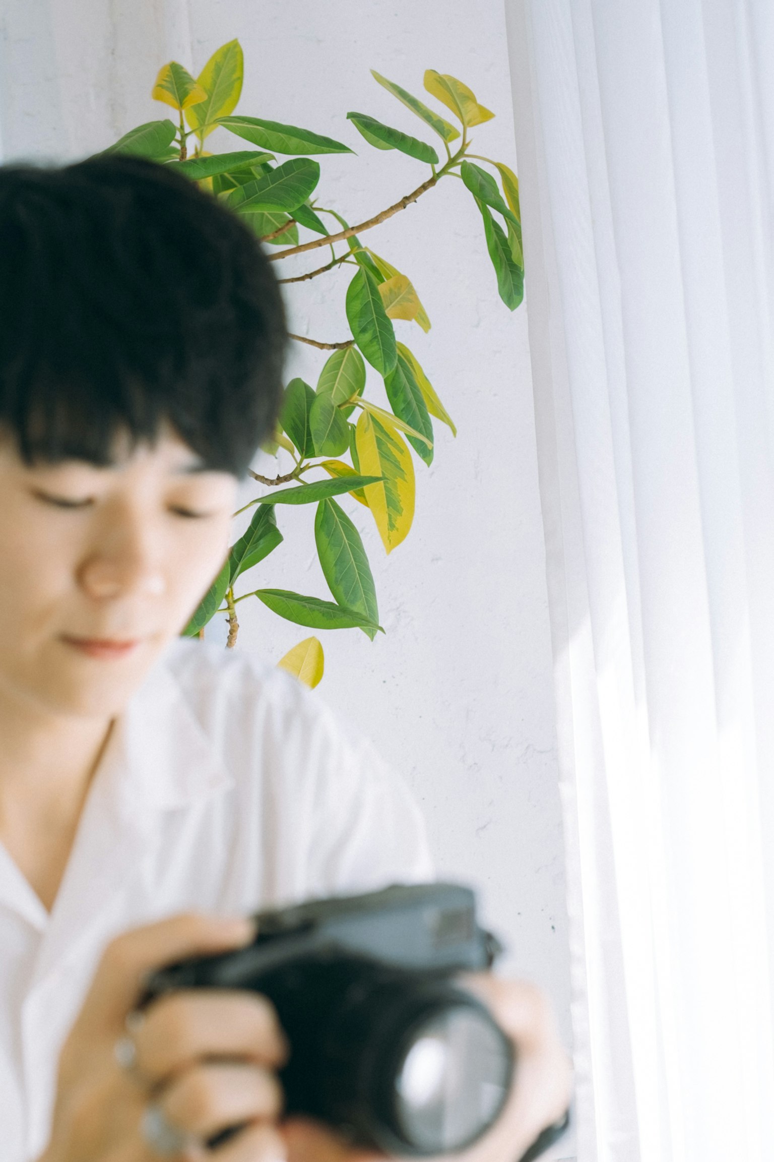 Young man holding a camera with green plant in the background indoors