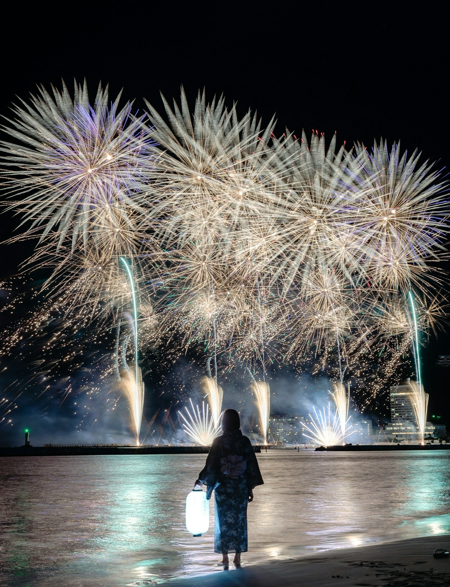 A person in a kimono holding a lantern watching fireworks in the night sky