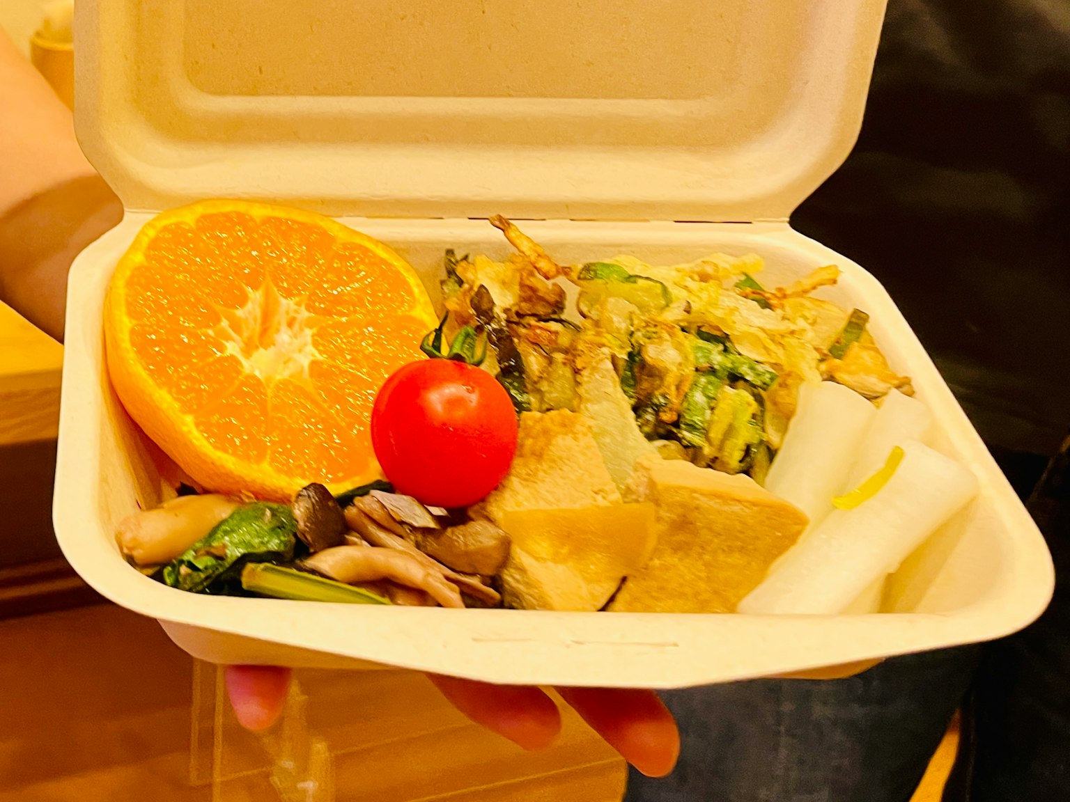 Una mano sosteniendo un recipiente de comida con una naranja, un tomate, verduras y tofu