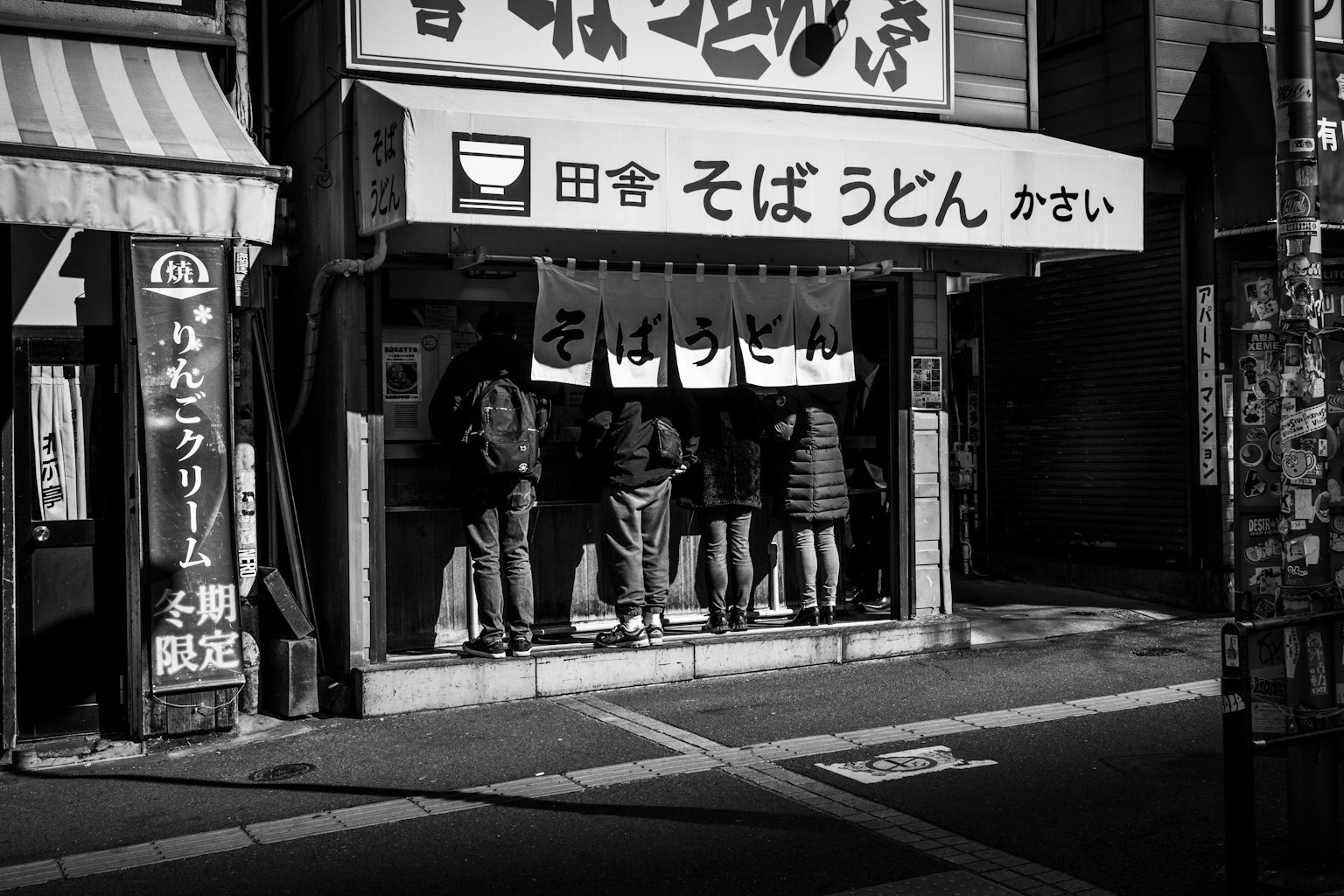 Personas esperando afuera de una tienda de soba udon en una esquina de la ciudad en blanco y negro