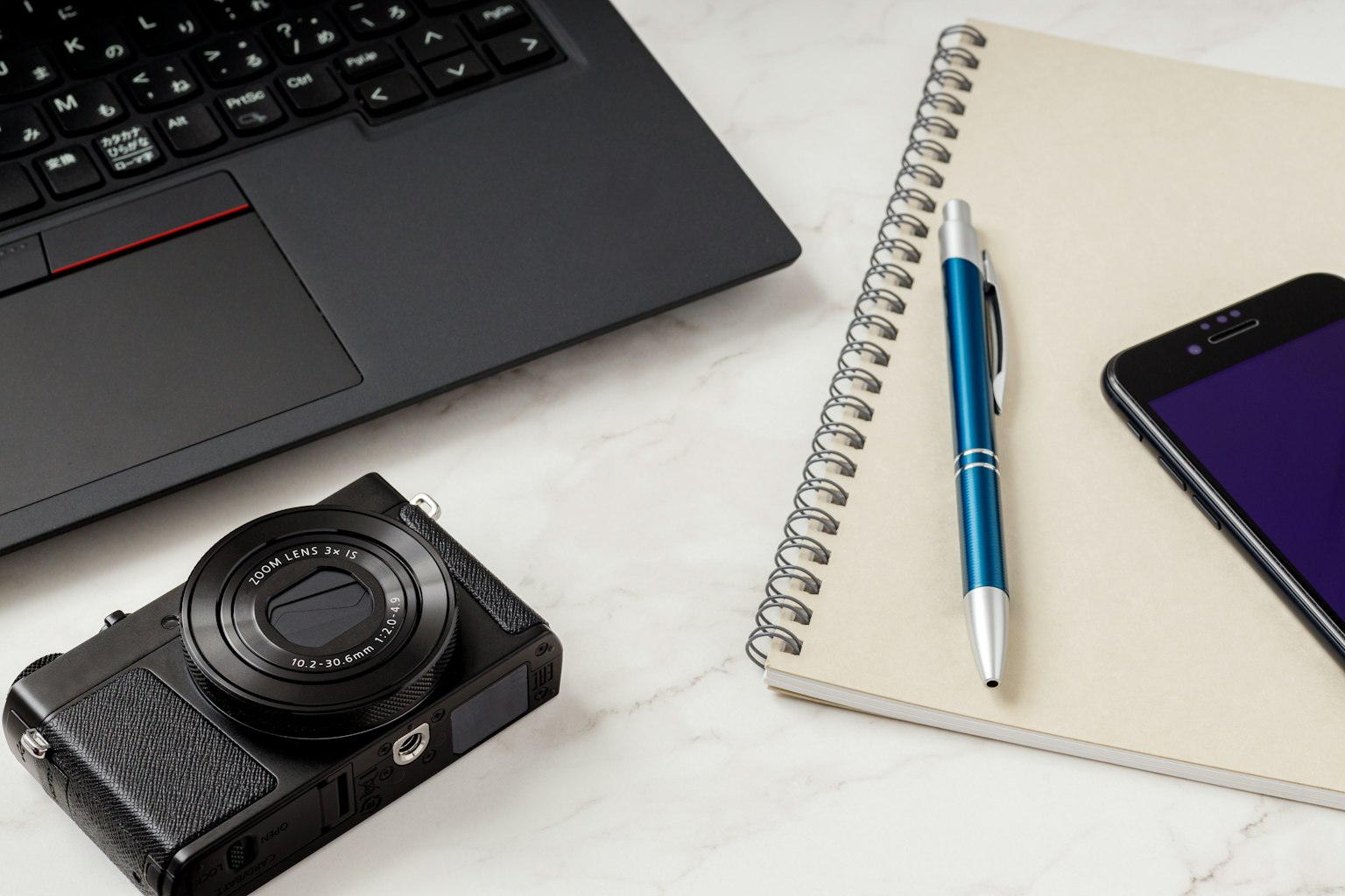 Una fotocamera, un laptop, una penna, un quaderno e uno smartphone disposti su una scrivania
