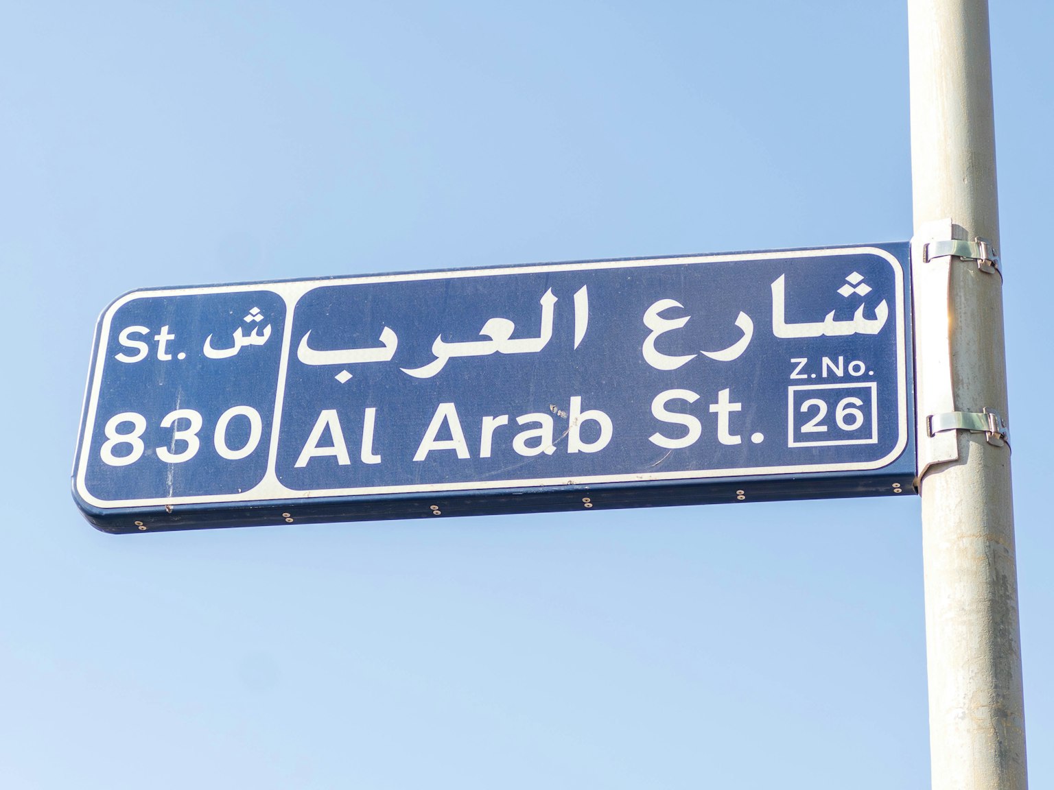 Blaues Straßenschild unter klarem Himmel mit Al Arab St 830 auf Arabisch und Englisch