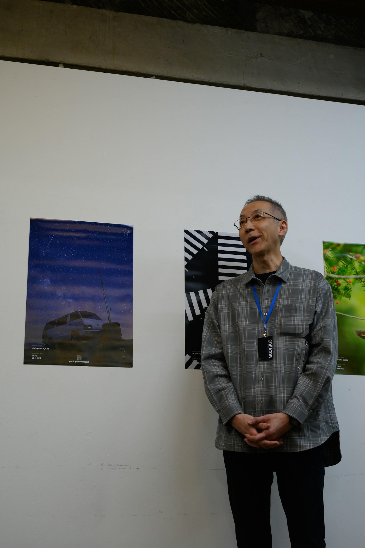 An elderly man in a gray shirt speaking at an exhibition with posters in the background