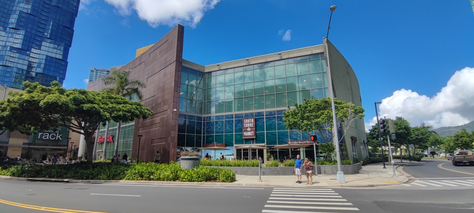Exterior de un edificio comercial moderno con fachada de vidrio y vegetación circundante bajo un cielo azul