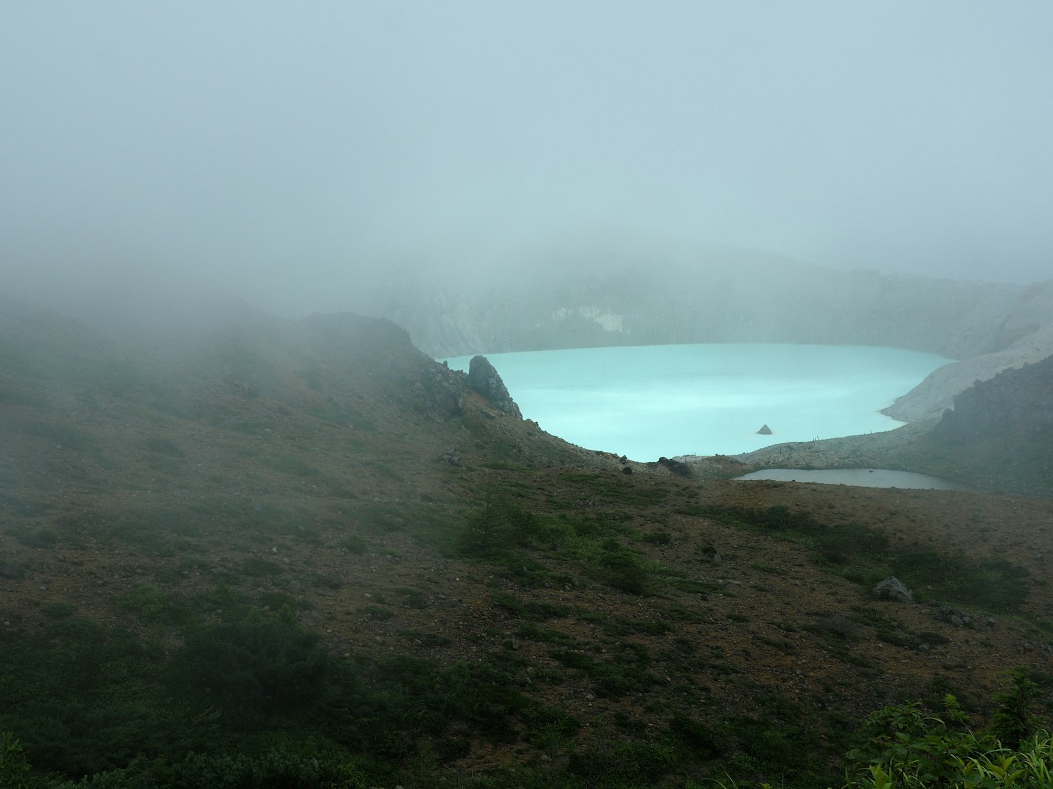 Pemandangan berkabut dari danau turquoise dikelilingi perbukitan hijau