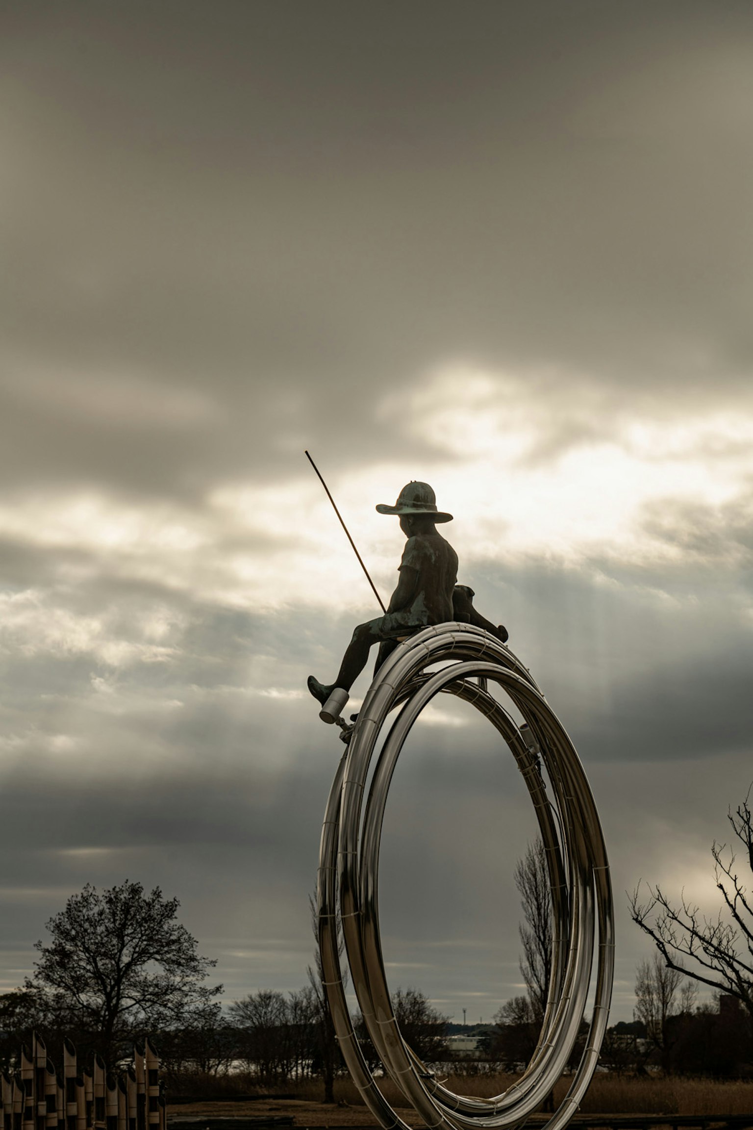 Skulptur eines Fischers, der auf einem silbernen Ring sitzt, umgeben von bewölktem Sonnenuntergang