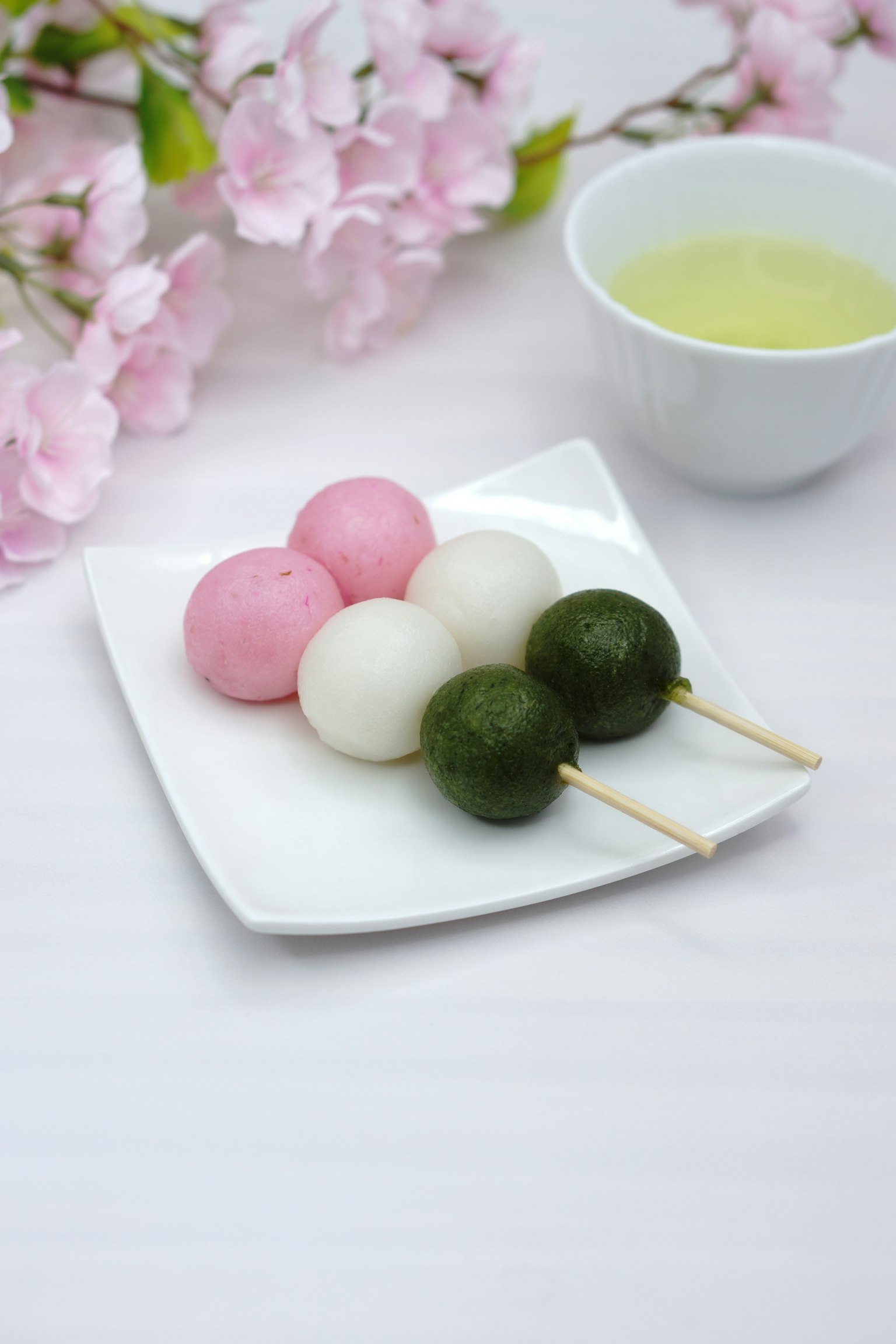 Dango colorido en un plato blanco con flores rosas y té verde al fondo