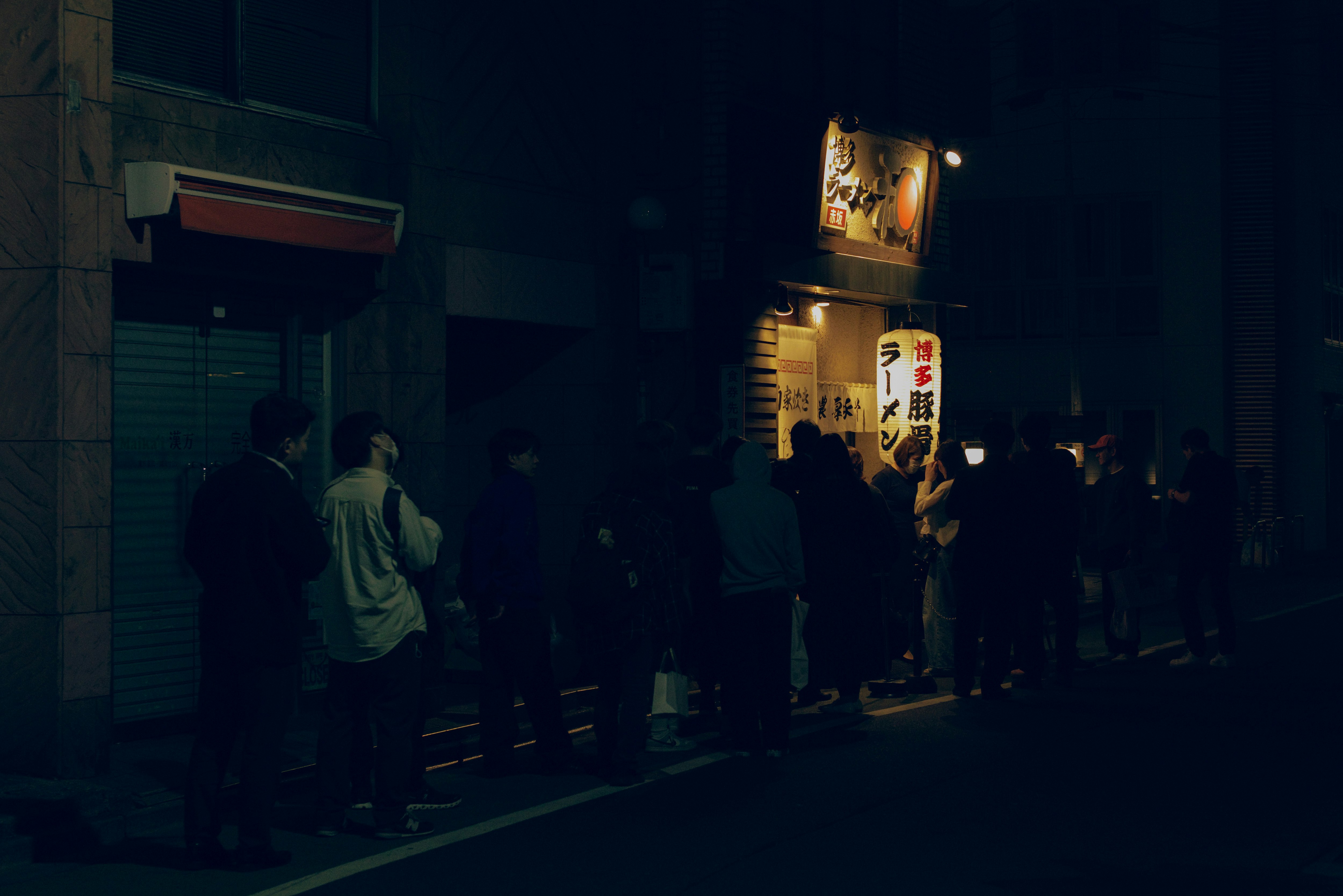 夜の街並みに並ぶ人々とラーメンの看板のある路地