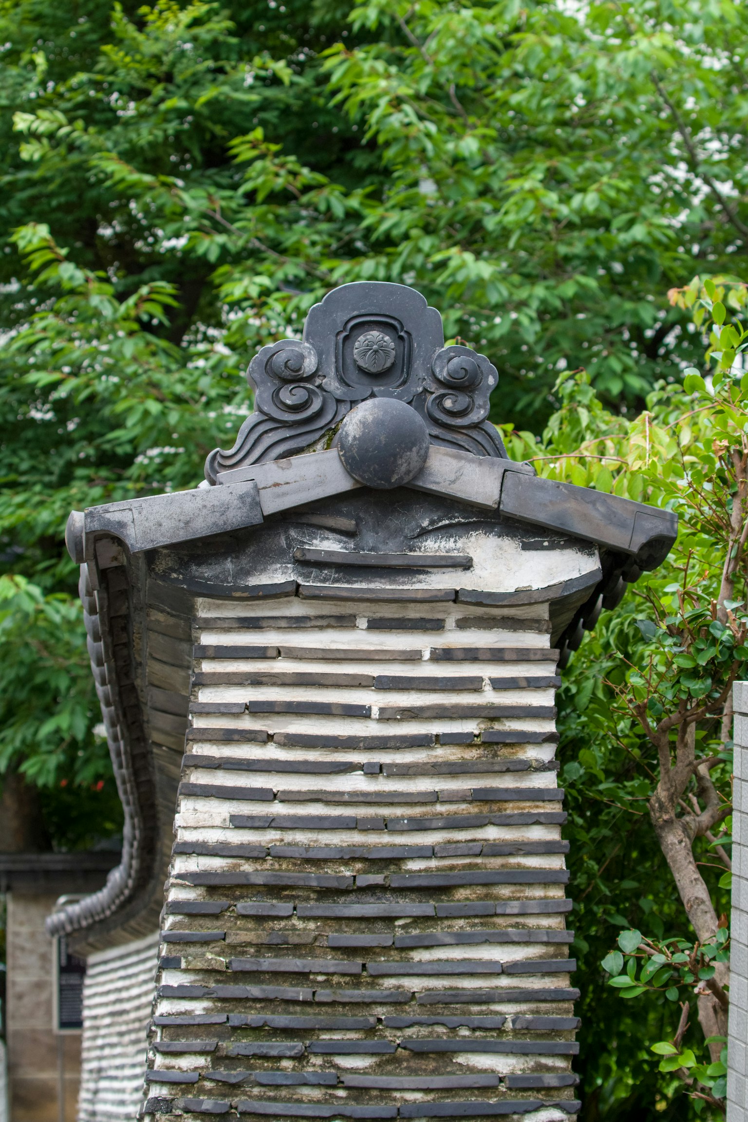 Part of an old building's roof featuring black tiles and stone walls with green trees in the background