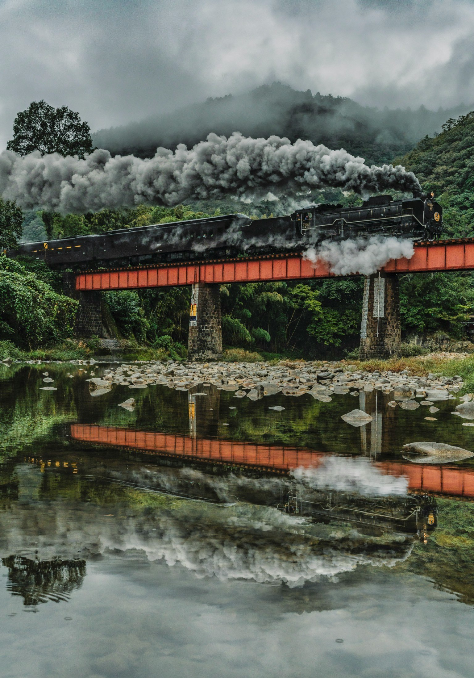 蒸気機関車が橋を渡る風景 濃い雲と山々の背景 水面に映る機関車の姿