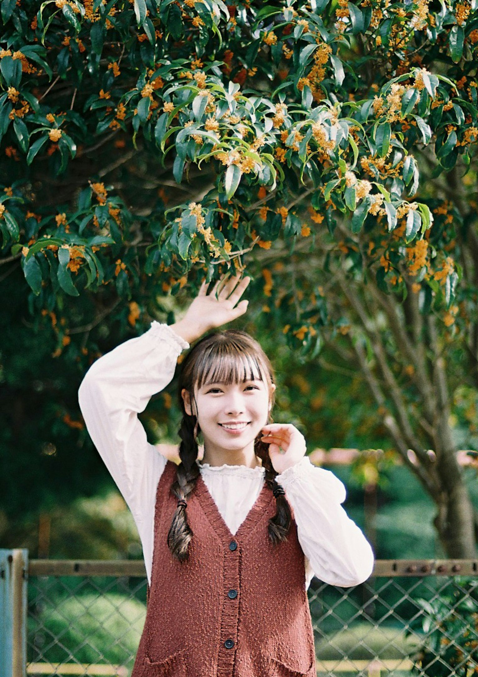 女孩在绿色树前摆姿势，穿着棕色背心和白色衬衫