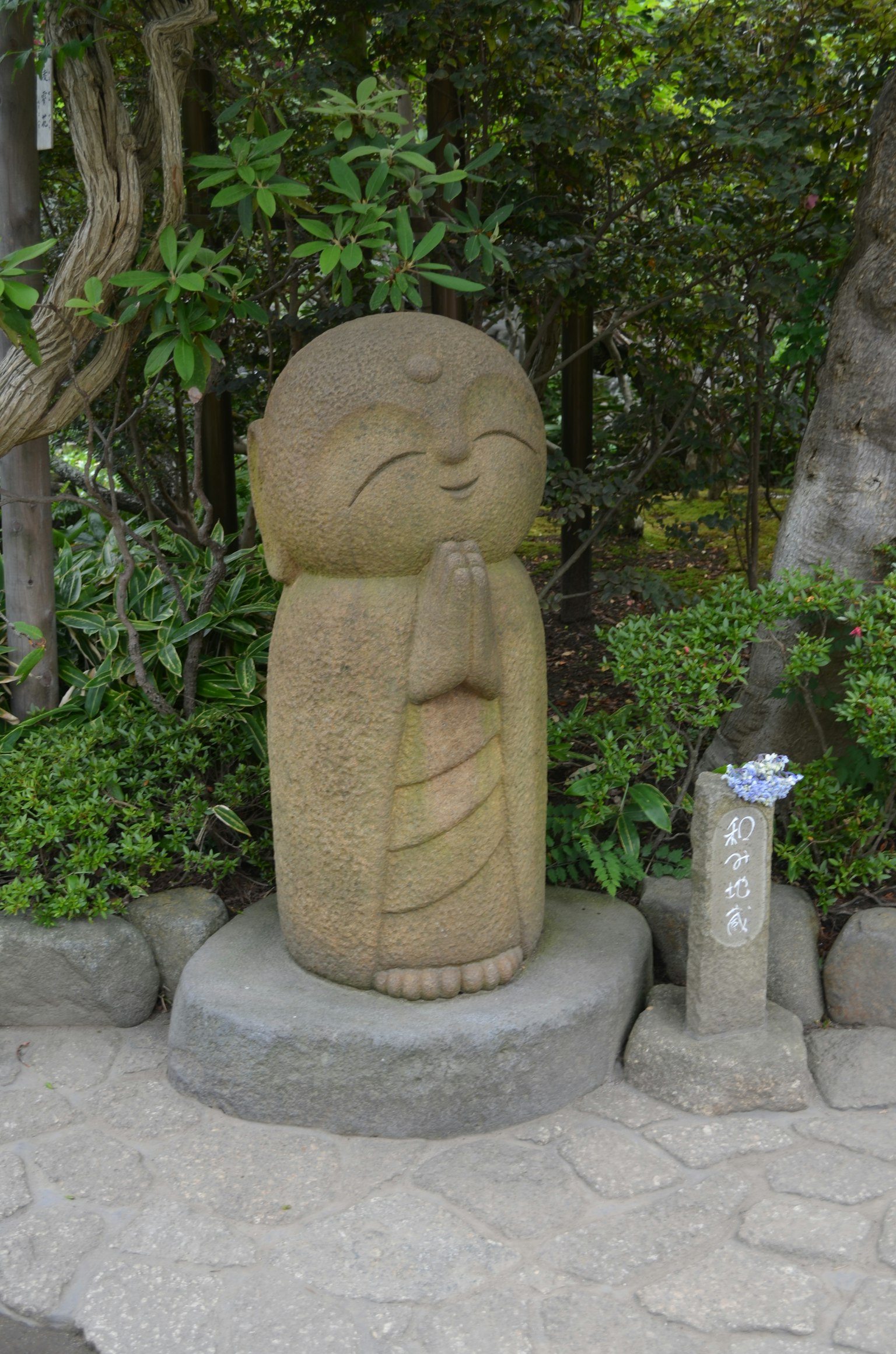 Stone statue of a Buddha figure with hands in prayer surrounded by green plants