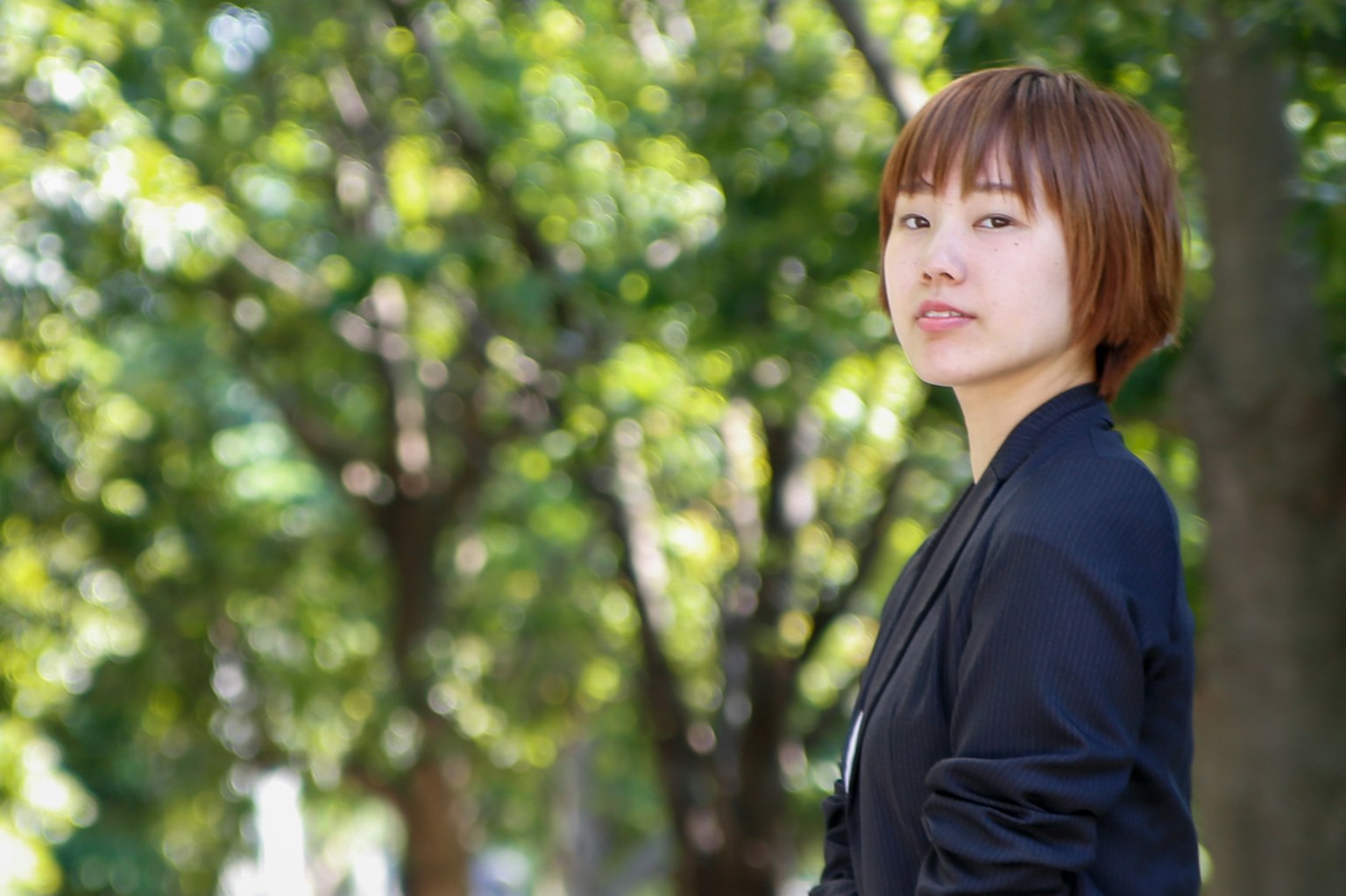 Portrait of a woman standing against a backdrop of green trees wearing a black jacket and short hair