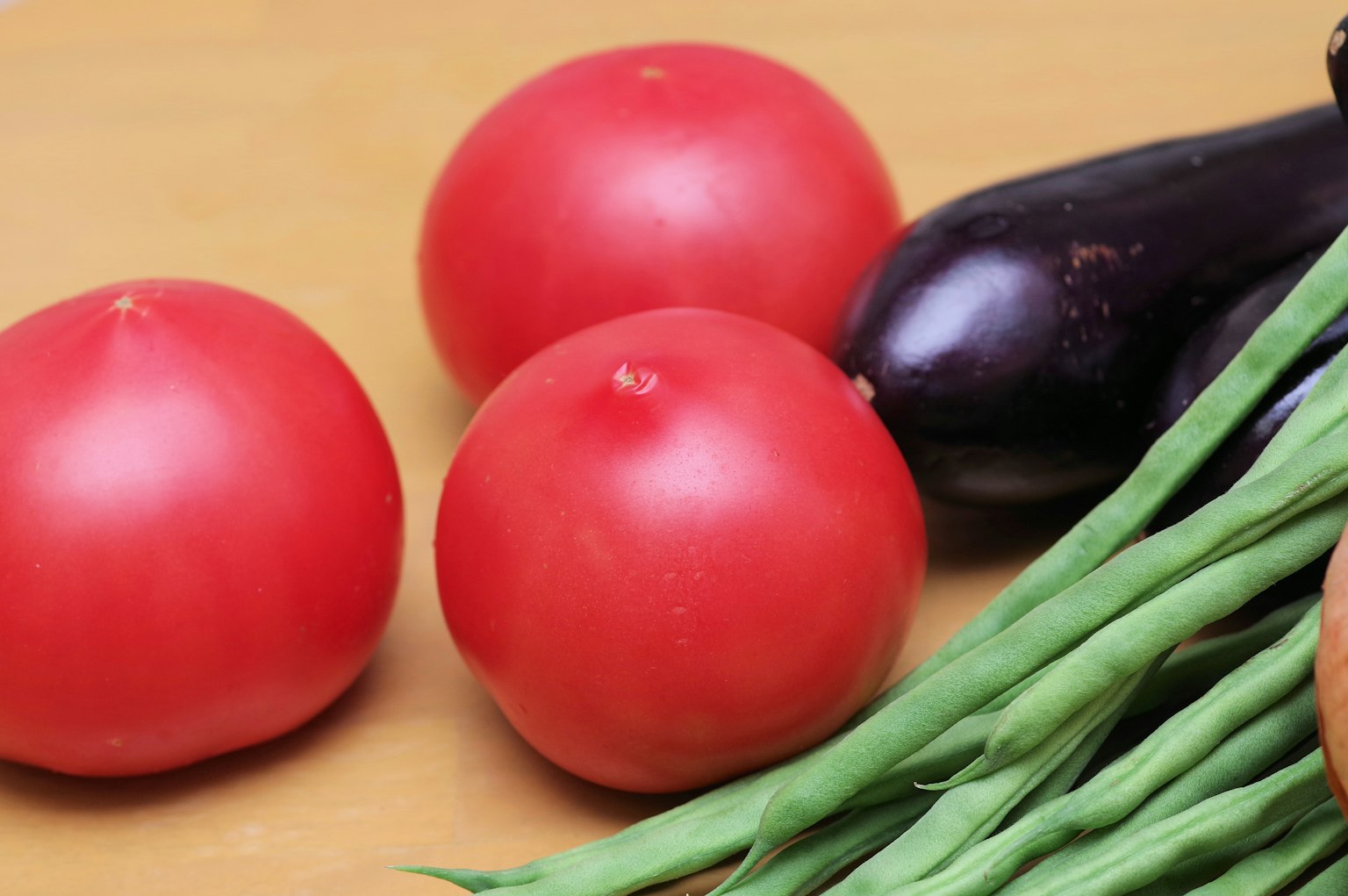 Drei rote Tomaten mit einer Aubergine und grünen Bohnen auf einer Holzoberfläche