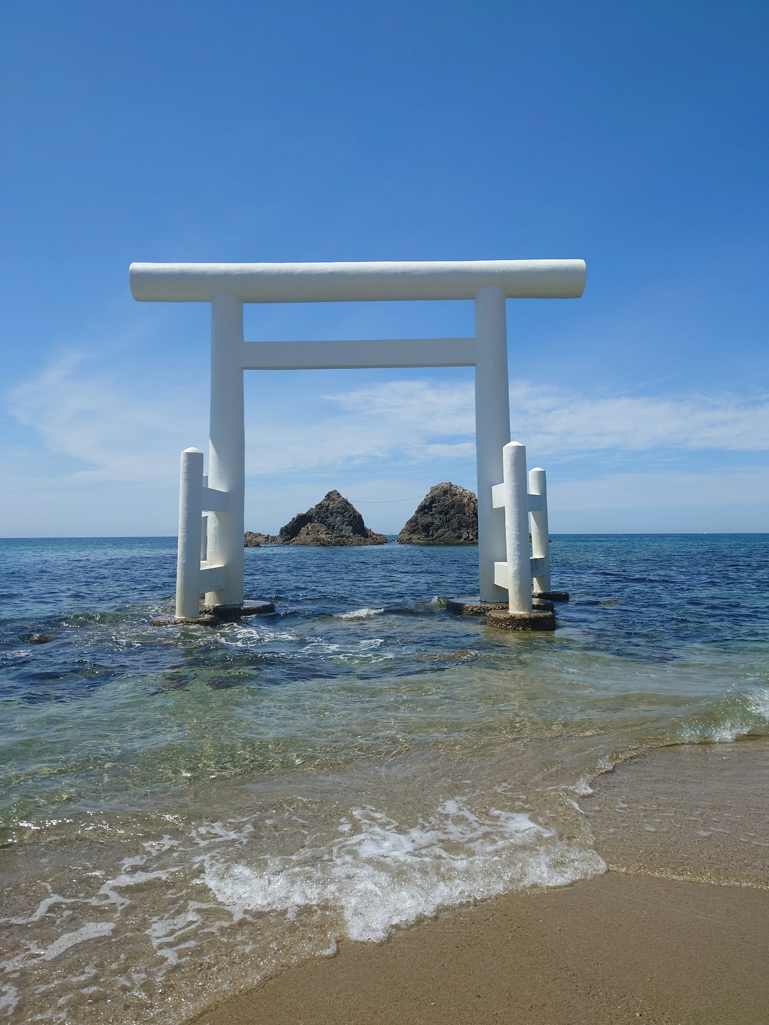 白色鳥居矗立在淺水中，背景是岩石和晴朗的藍天