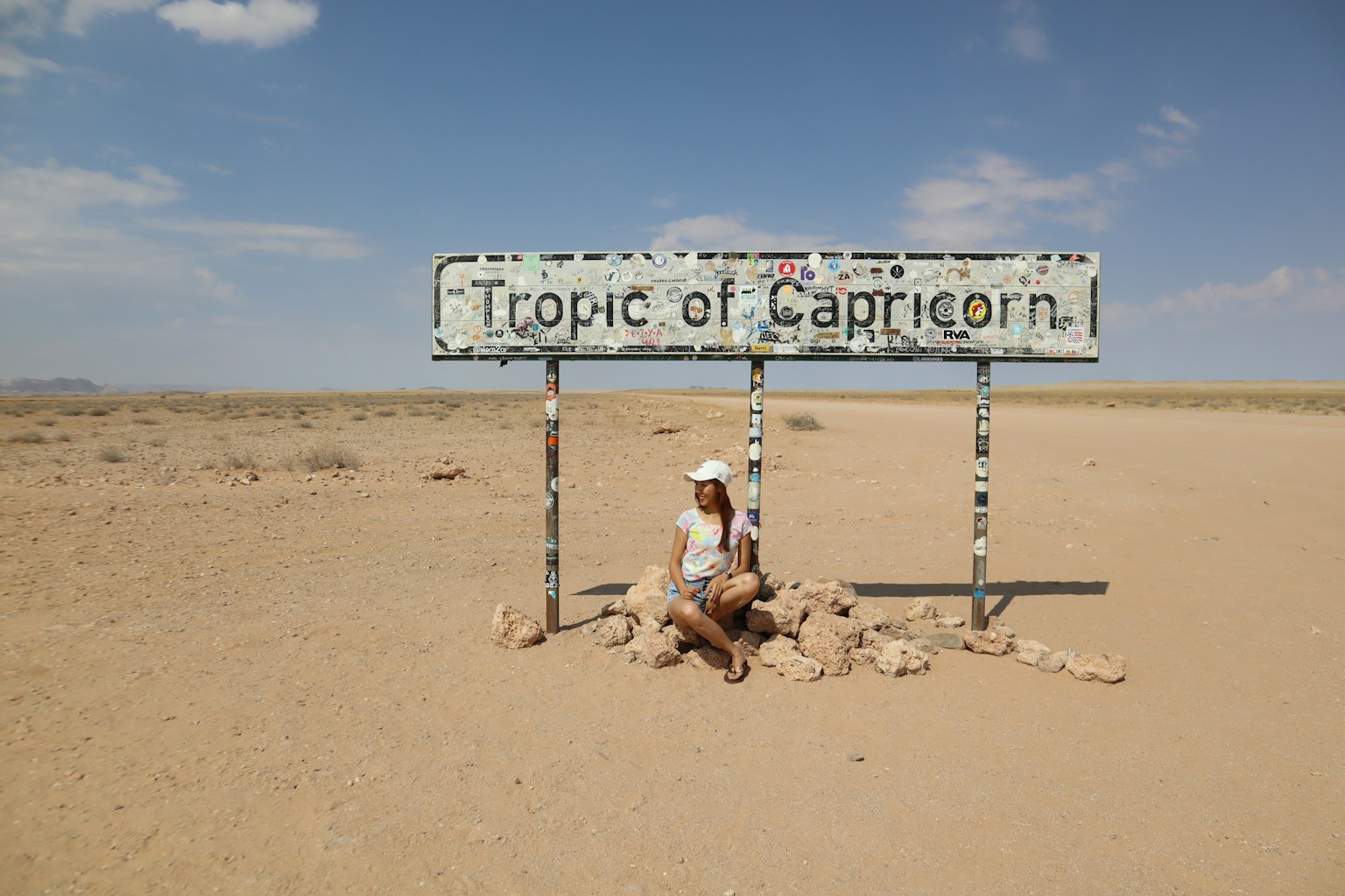Wanita duduk di depan tanda Tropic of Capricorn di lanskap gurun dengan langit biru