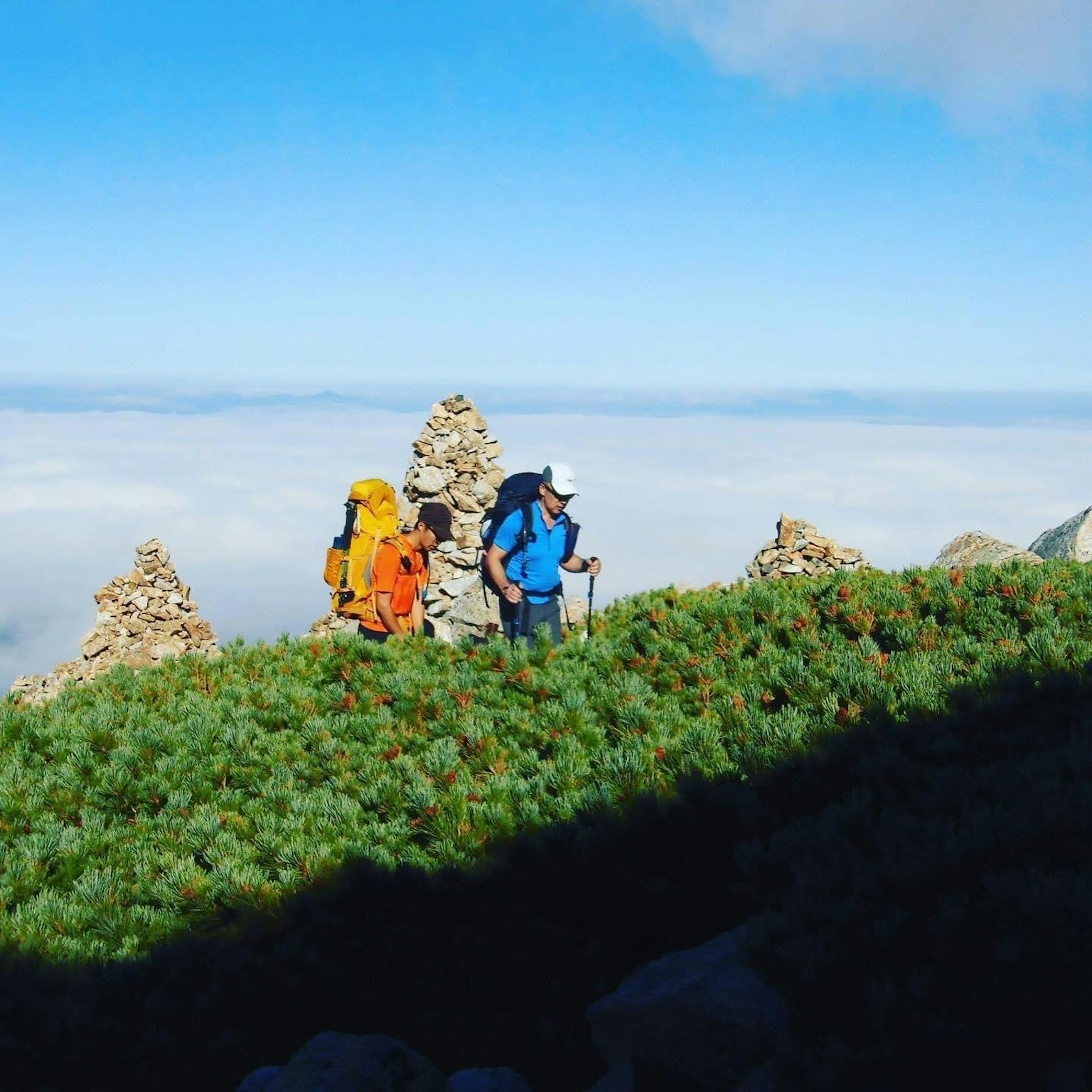 Dos excursionistas en la cima de una montaña con estructuras de piedra y un mar de nubes