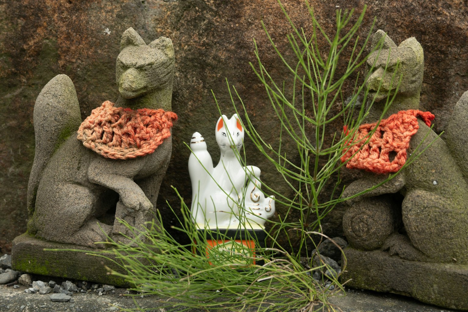 Zwei Steinfuchs-Statuen mit gestrickten Schals und einer weißen Katzenfigur in der Mitte, umgeben von Grün