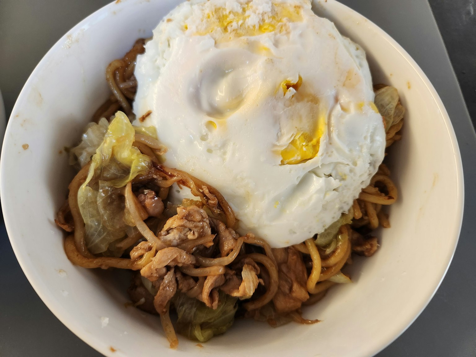 一碗炒麵，上面放著一顆煎蛋和混合蔬菜