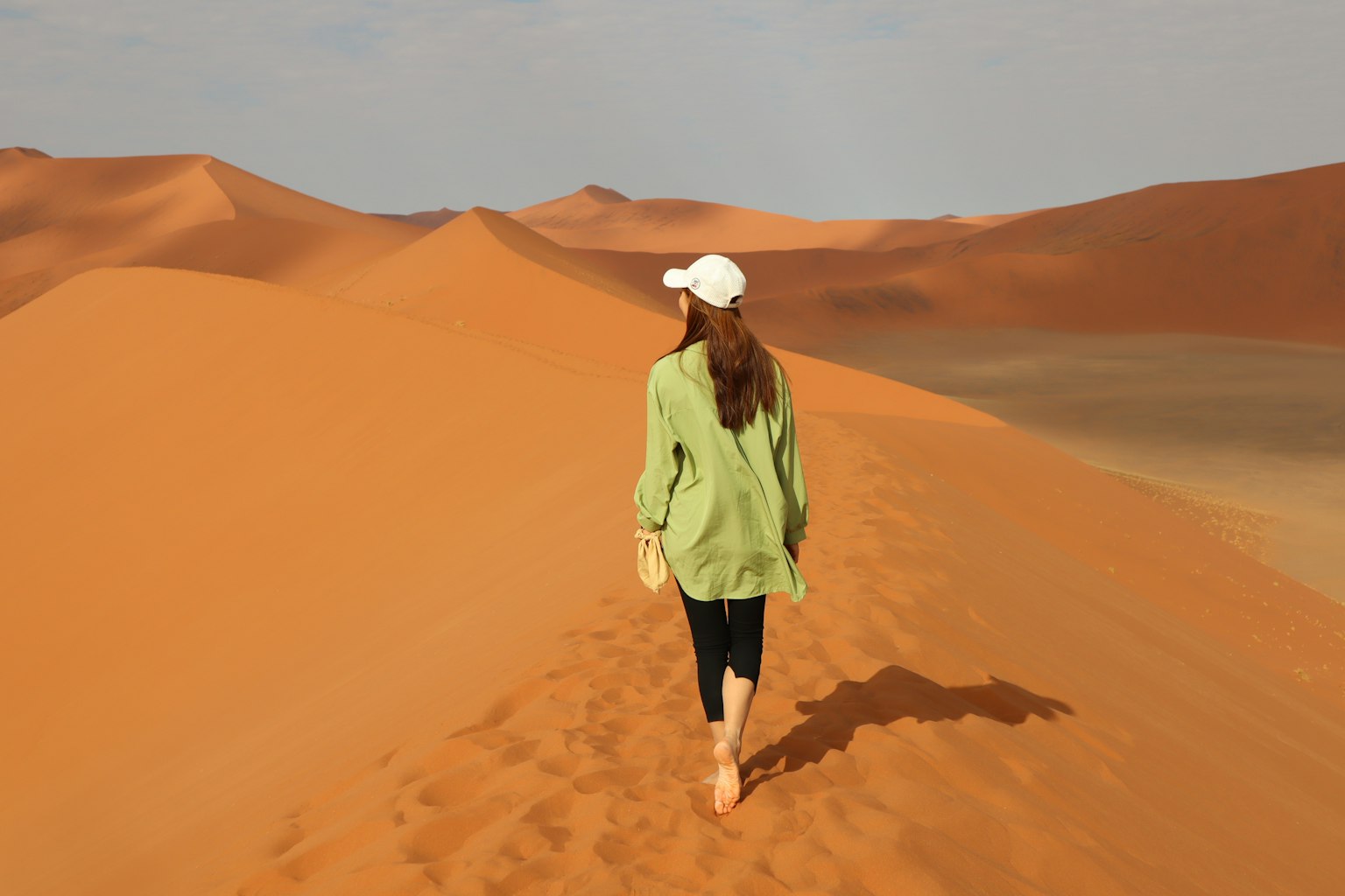 Mujer caminando sobre dunas de arena con una camisa verde y un sombrero blanco