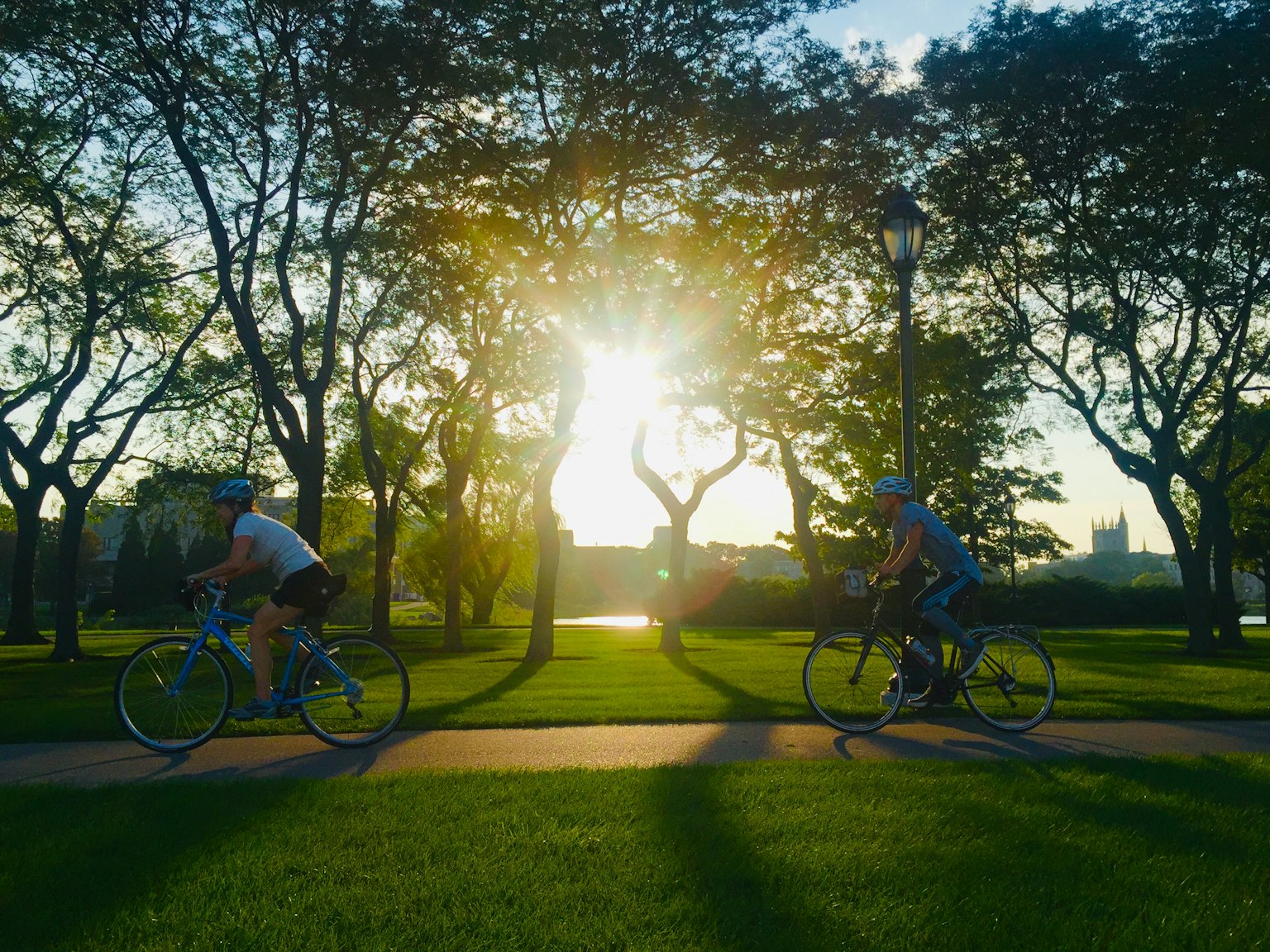 Two cyclists riding along a path in a park with the sunrise in the background