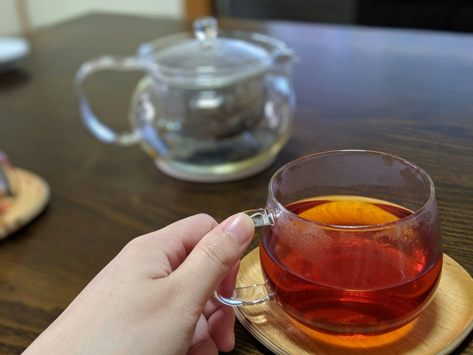 Eine Hand hält eine transparente Teetasse mit rotem Tee und eine Teekanne im Hintergrund