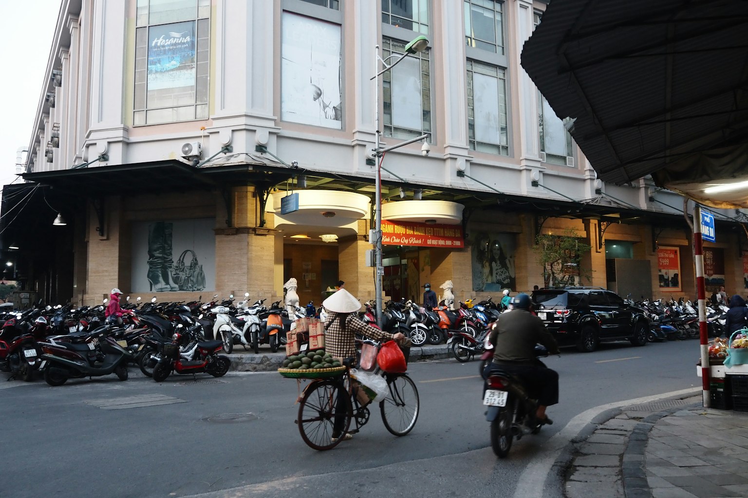 街角場景，自行車和摩托車經過，人們在建築物前聚集