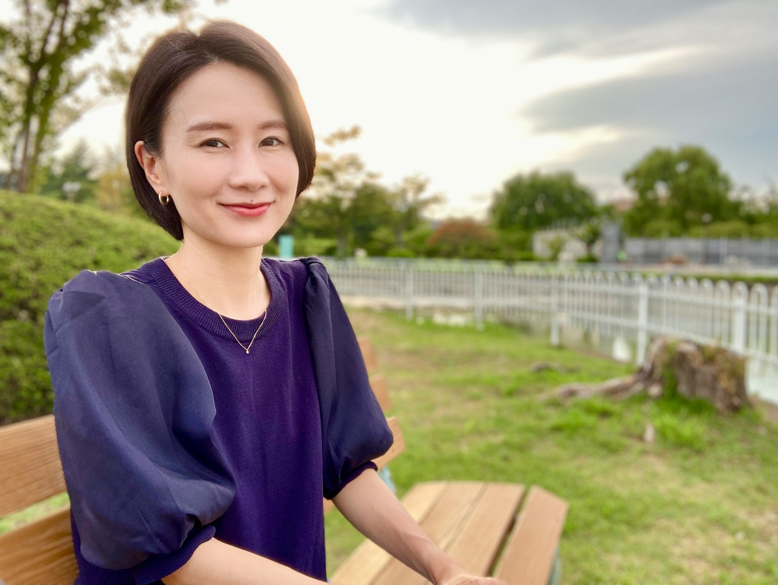 ภาพพอร์ตเทรตของผู้หญิงนั่งอยู่บนม้านั่งในสวน ยิ้มและมองไปที่กล้อง สวมเสื้อสีม่วง