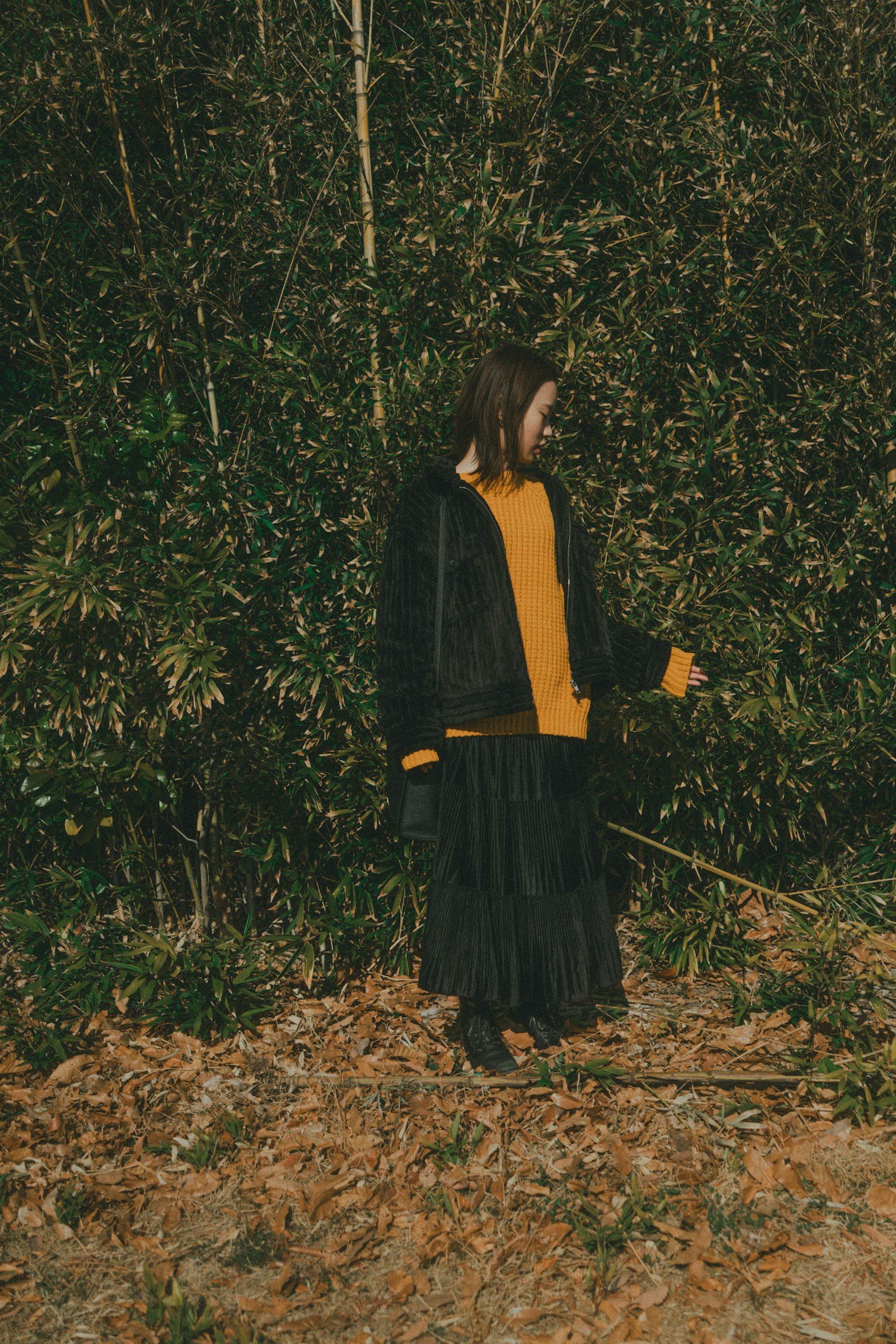 Seorang wanita mengenakan pakaian hitam dan sweater kuning berdiri di hutan