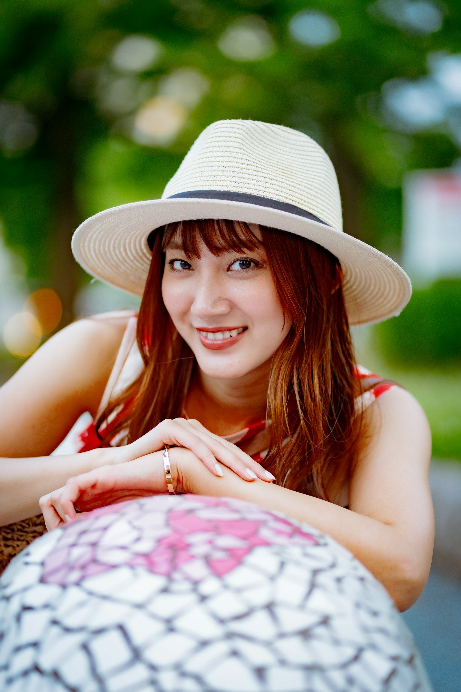 Eine lächelnde Frau mit einem weißen Hut und einem bunten Kleid, die im Freien sitzt, umgeben von Grün