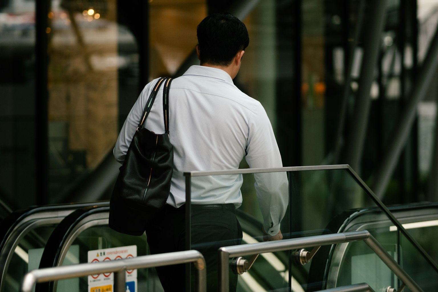 Man in white shirt ascending escalator carrying black bag