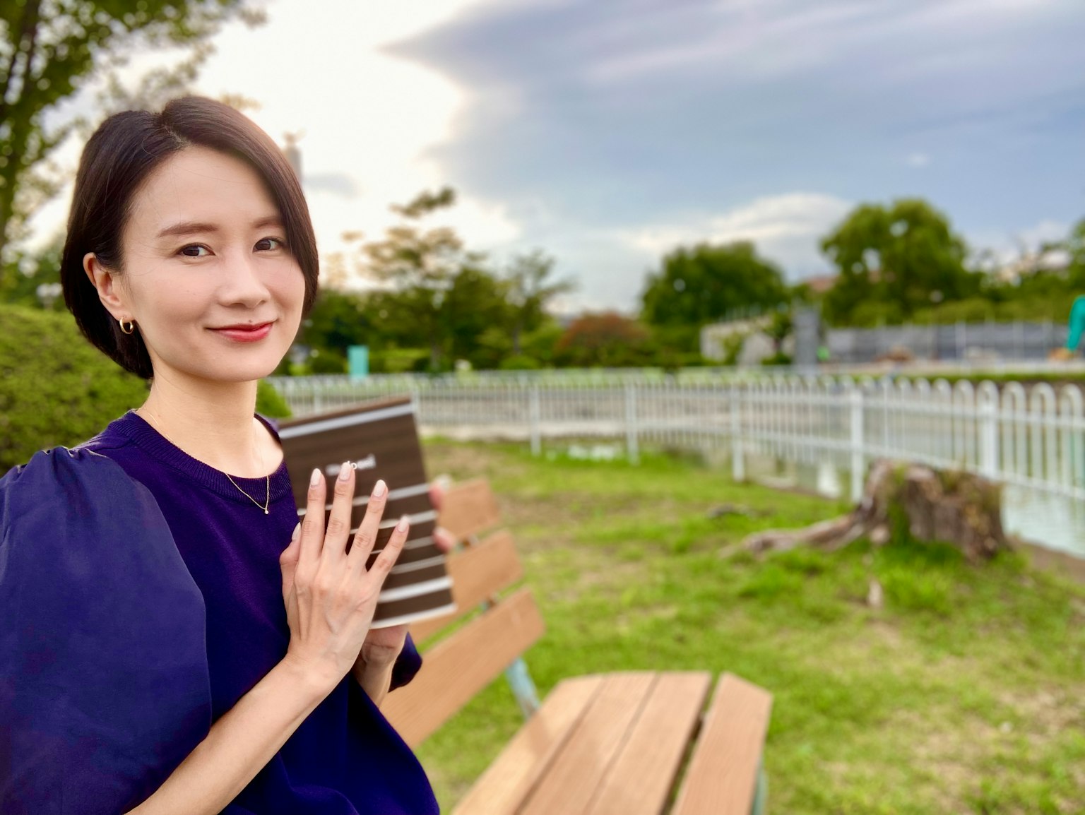 ผู้หญิงนั่งอยู่บนม้านั่งในสวนถือหนังสือและยิ้ม