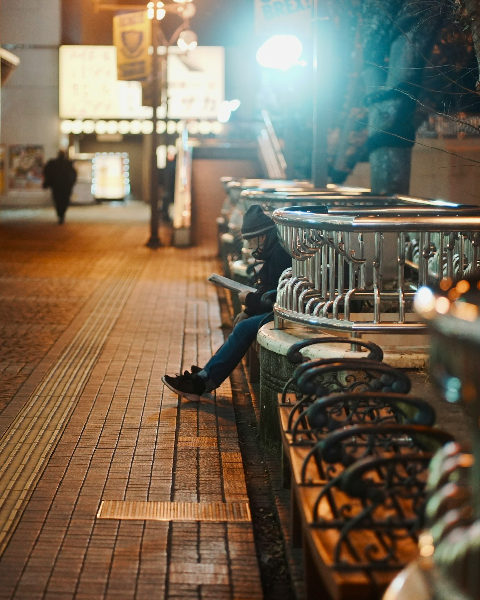 Una persona seduta su una panchina di notte con un'altra che cammina per strada