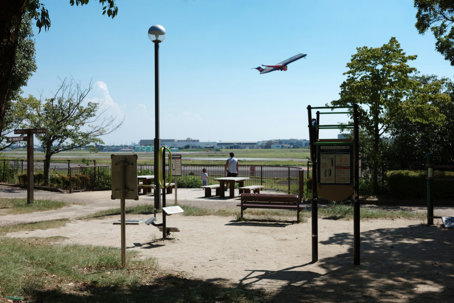 公園のベンチと遊具があり、空を飛ぶ飛行機が見える