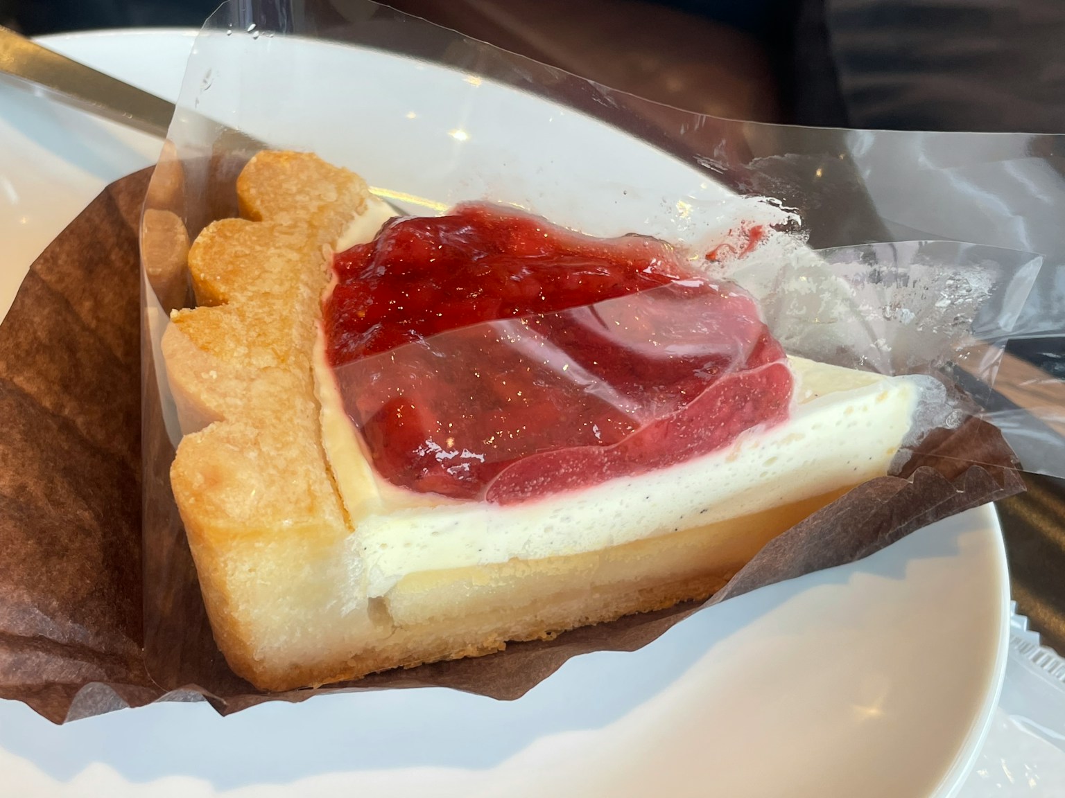 Una fetta di cheesecake guarnita con salsa di fragole avvolta in plastica trasparente su un piatto bianco