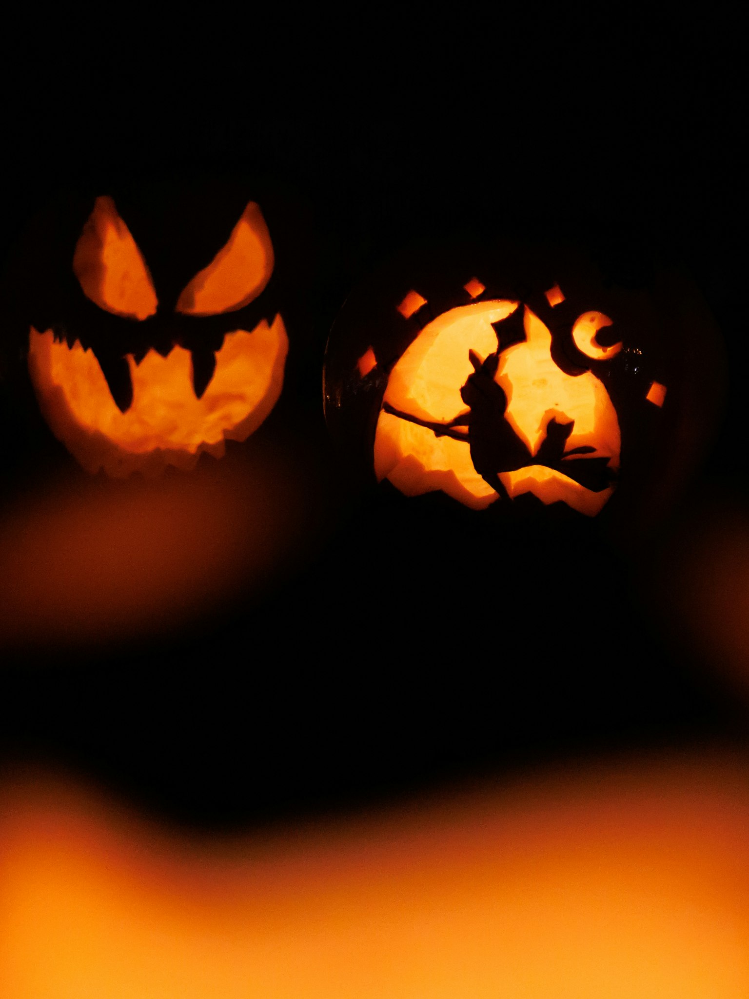 ハロウィンのカボチャの彫刻が二つ並んでいる一枚の画像 一つは怖い顔のデザインでもう一つは魔女が月の前を飛んでいるデザイン