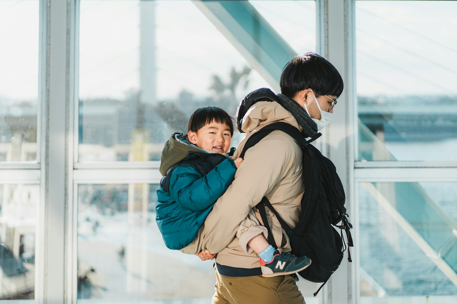 A parent carrying a child in a backpack near large windows with a view of a harbor