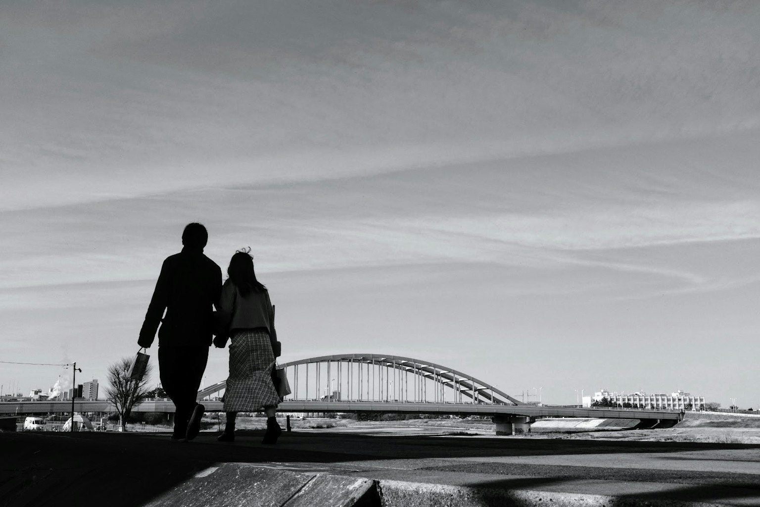 Silhouette hitam putih pasangan berjalan bergandeng tangan dengan jembatan lengkung di latar belakang