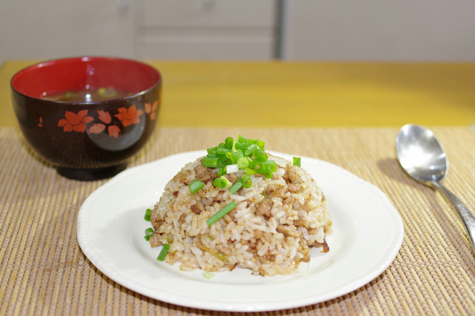 Fried rice topped with green onions on a white plate with a red bowl of soup