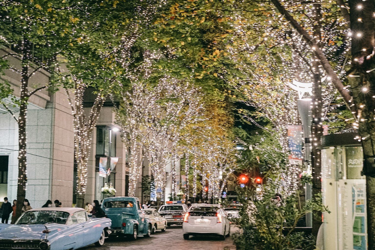 Una calle animada adornada con luces brillantes en los árboles y coches antiguos estacionados a lo largo de la carretera