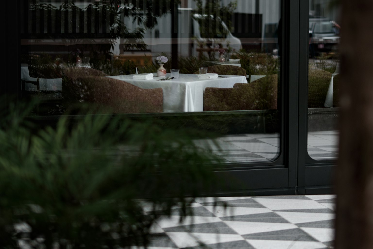 Mesa de restaurante con mantel blanco vista a través de una ventana plantas verdes y baldosas monocromáticas en primer plano