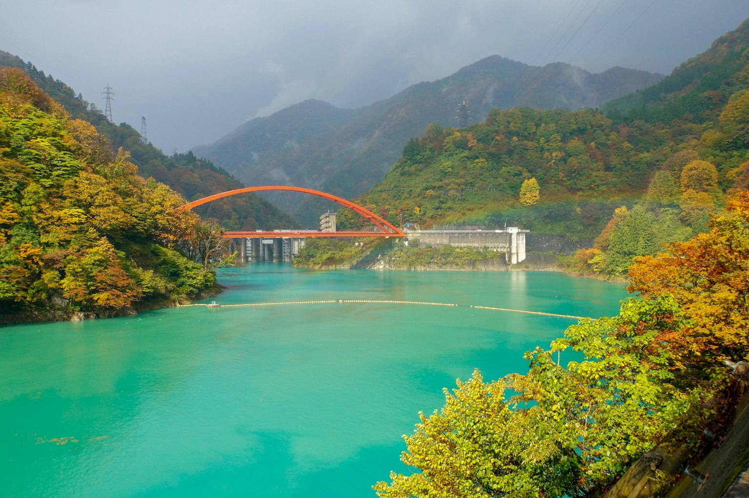 ทะเลสาบสีฟ้าอมเขียวที่สวยงามล้อมรอบด้วยภูเขาสีฤดูใบไม้ร่วงพร้อมสะพานโค้งสีแดง