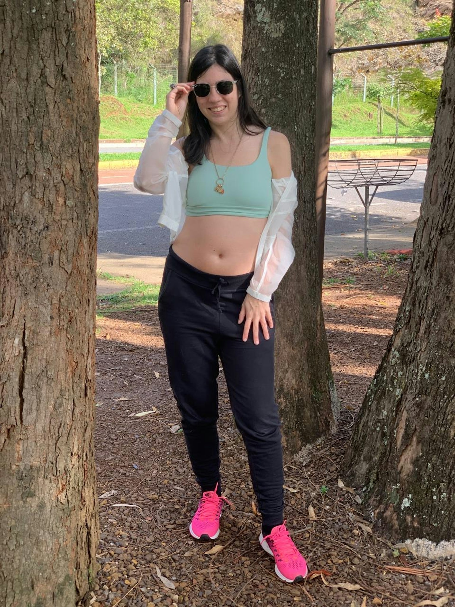 Mujer de pie entre árboles usando una blusa azul claro y pantalones negros con zapatillas rosas y gafas de sol
