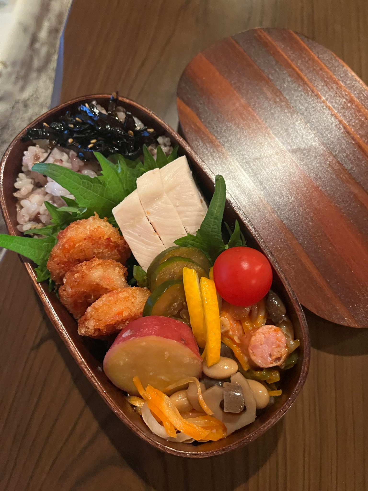 Une boîte à bento joliment disposée avec des plats colorés comprenant des crevettes frites, du tofu, des légumes, des tomates cerises et des fruits