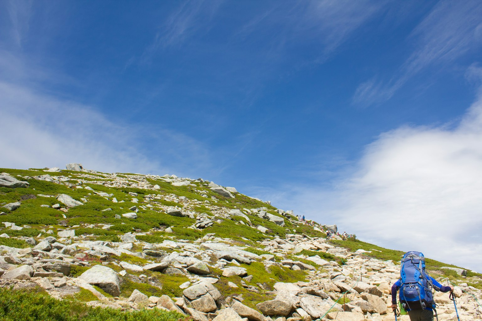Hiker with a large backpack climbing a rocky hill under a blue sky