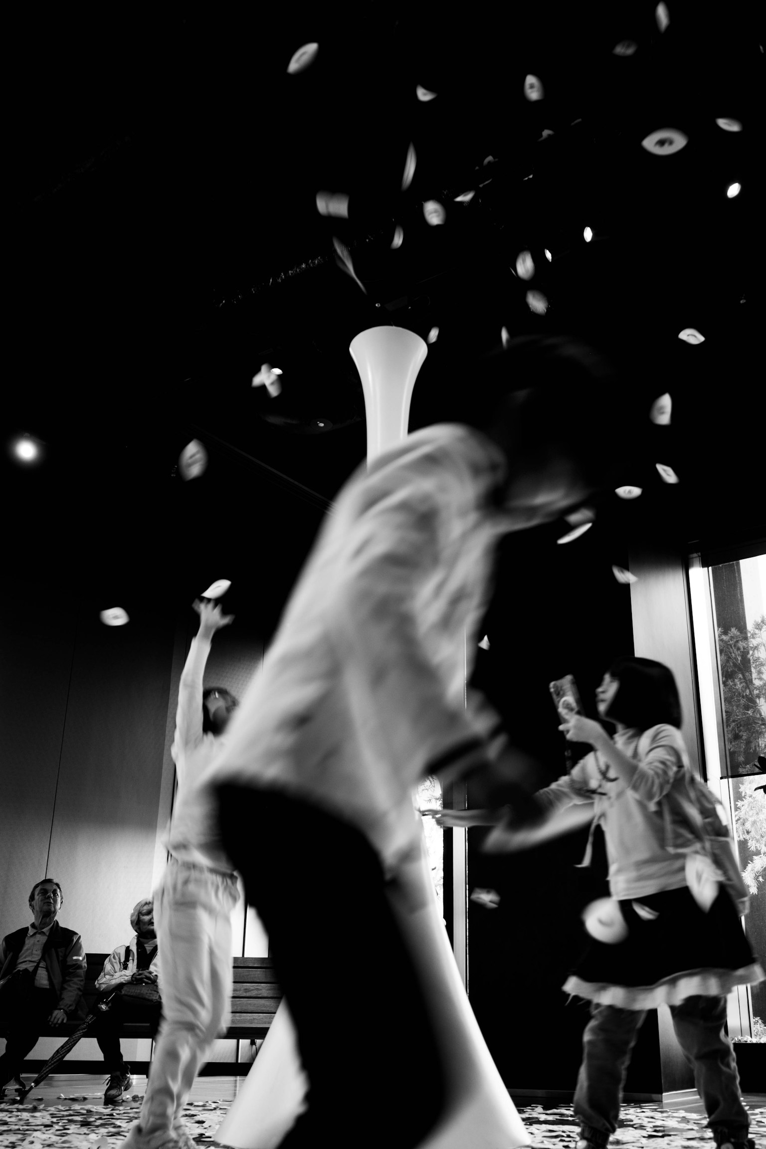 Black and white photo of children playing joyfully. A white column stands in the center with colorful objects floating around. Background features seated spectators.